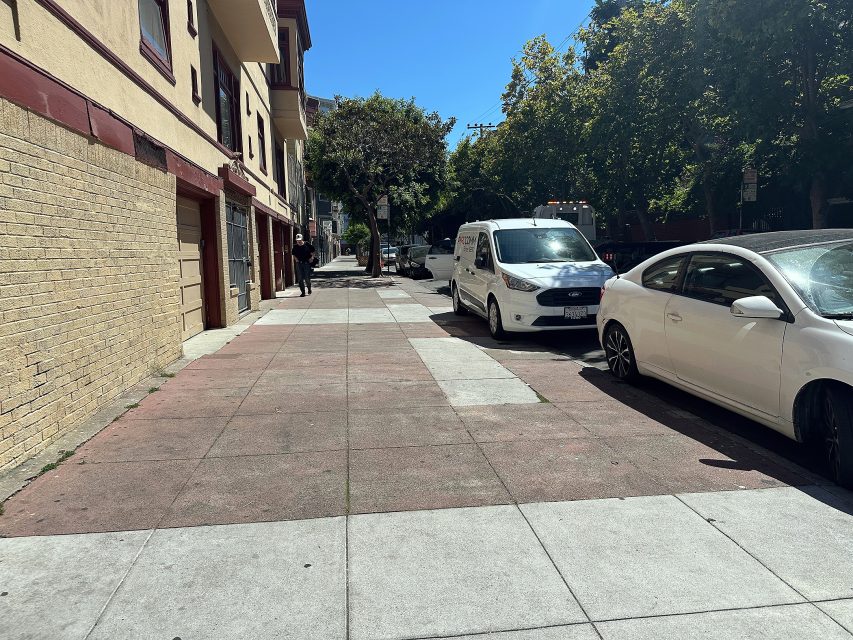 Sunny urban sidewalk with parked white cars along the curb, apartment buildings on the left, and trees lining the street.