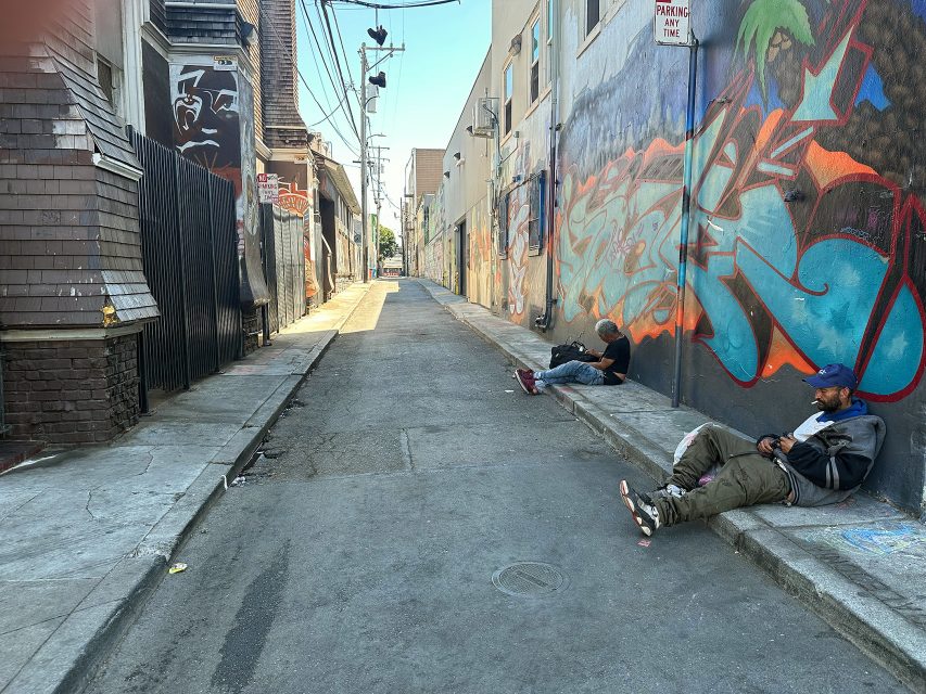 Two men sit on the sidewalk in a narrow alley lined with buildings and colorful graffiti during daylight.