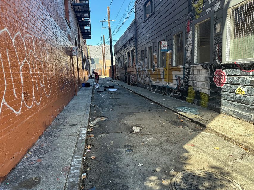 A narrow urban alley with graffiti on the walls, scattered trash on the ground, and a person bending over near the center.