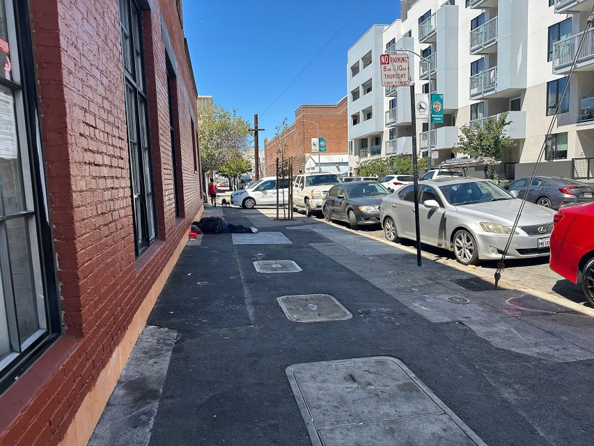 A city sidewalk next to a red brick building with parked cars along the street and a no parking sign visible. A person lies on the sidewalk in the distance under a blanket.