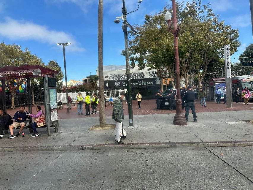 People are gathered at a city street corner near a bus stop; some are sitting, others standing, with police officers and utility workers present. Trees and buildings are in the background.