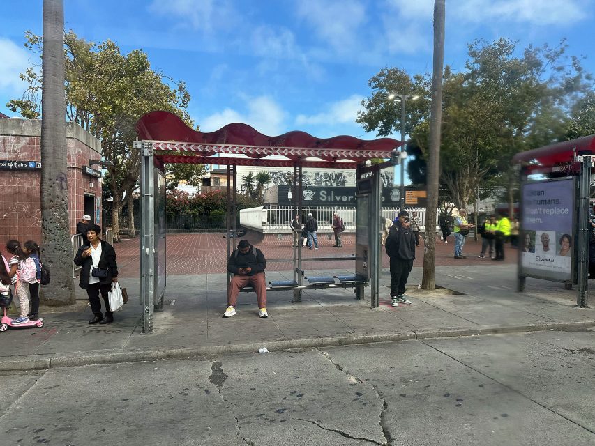A man sits at a bus stop shelter while other people stand and wait nearby; trees and buildings are visible in the background.