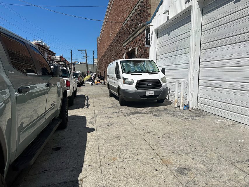 A white van is parked in front of a garage door on a sunny day, with other vehicles and people visible further down the sidewalk.