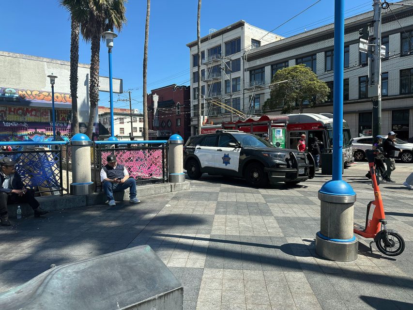 A police SUV is parked in a city plaza near people sitting on benches, with a red streetcar and buildings in the background on a sunny day.