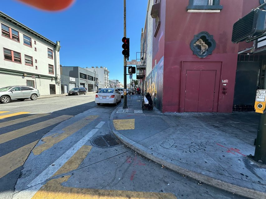 Urban street scene with parked cars, pedestrians on the sidewalk, a red building, and a traffic light under a clear blue sky.