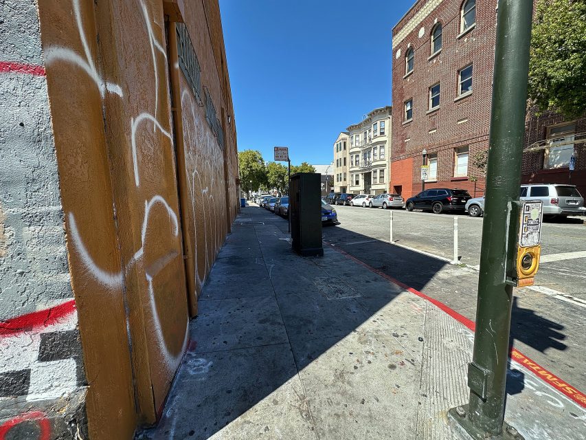 A city sidewalk with graffiti on a building wall, parked cars along the street, and a crosswalk button on a pole under a clear blue sky.