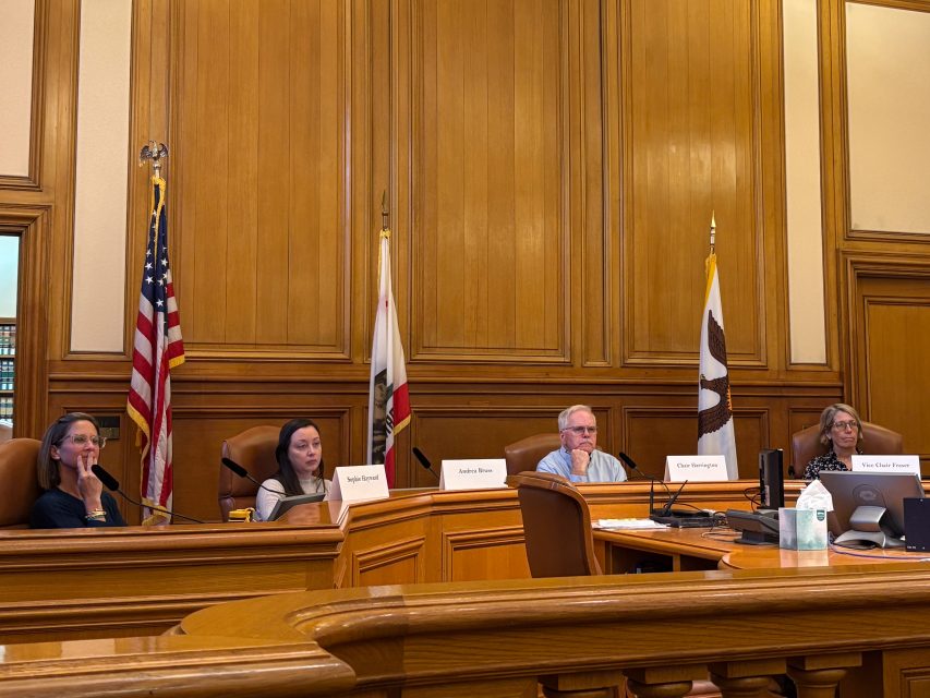 Four people sit at a wood-paneled council chamber desk with nameplates, microphones, and flags of the United States and California in the background.