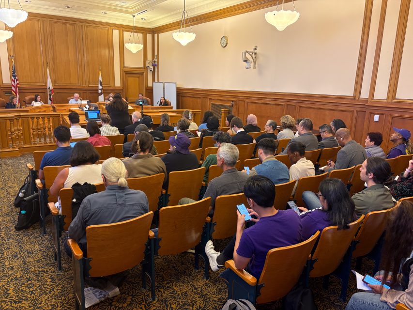 A group of people seated in a wood-paneled room attend a public meeting or hearing, with officials seated at a raised desk in front.