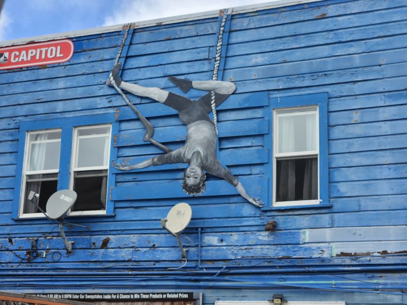A mural of a person in grayscale appears to hang upside down from a rope between two windows on a weathered blue wooden building with satellite dishes.