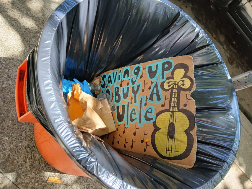 A cardboard sign reading "Saving up to buy a ukulele" with a drawing of a ukulele is discarded in a trash can with other garbage.