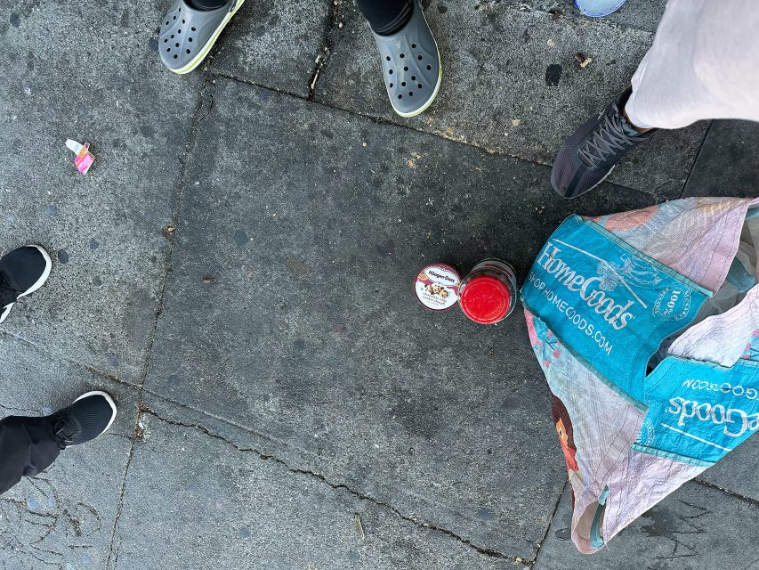 Four people stand on a sidewalk around an open jar of pasta sauce and a HomeGoods shopping bag. Pieces of trash are visible on the ground.