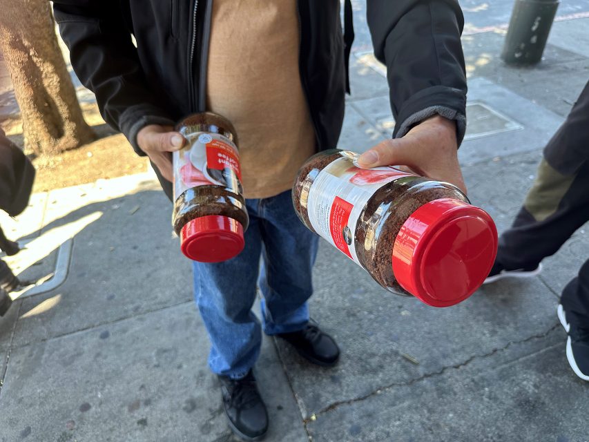 A person standing on a sidewalk holds two large jars of instant coffee, one in each hand.