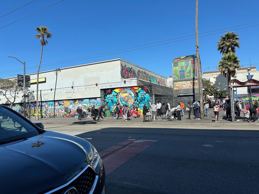 People stand in a line on a city sidewalk in front of a building covered with colorful graffiti murals on a sunny day. Cars are parked along the street.