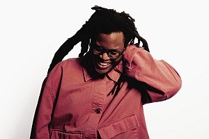 Person with long dreadlocks, wearing glasses and a loose-fitting, buttoned pink jacket, looking down and smiling with one hand behind their head, against a plain white background.