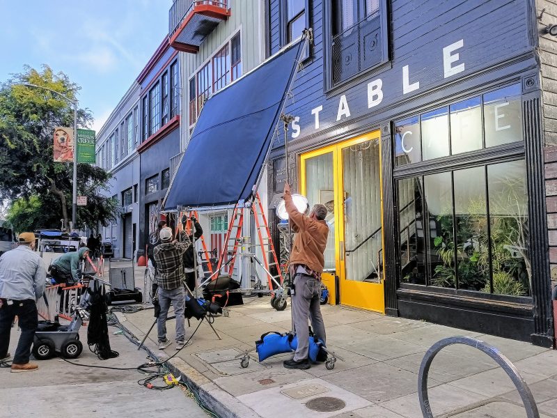 Film crew sets up lighting and equipment outside the entrance of Stable Cafe on a city street. Several people adjust a large blue screen and other gear on the sidewalk.