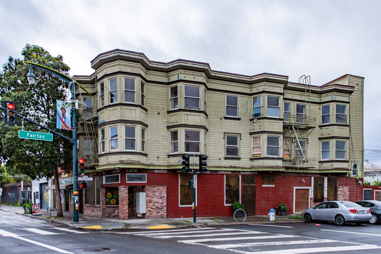 A three-story corner building with bay windows and ground-floor shops at a street intersection, with a gray car parked nearby and a traffic light visible.