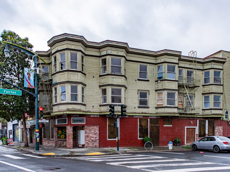 A three-story corner building with bay windows and ground-floor shops at a street intersection, with a gray car parked nearby and a traffic light visible.