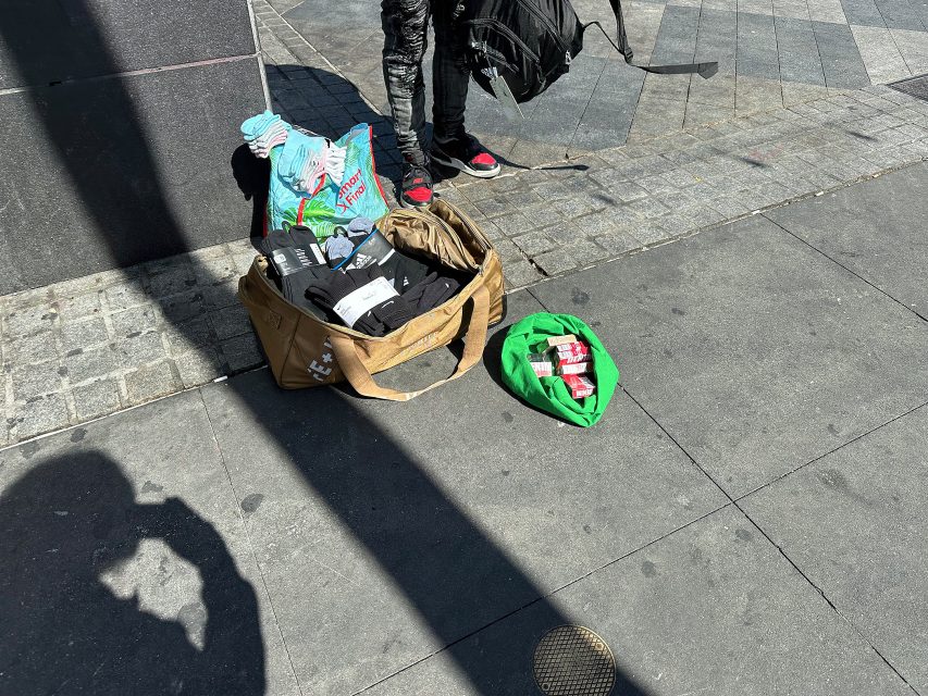 A duffel bag with clothes, a green bag with canned drinks, and a person standing nearby on a city sidewalk in sunlight.