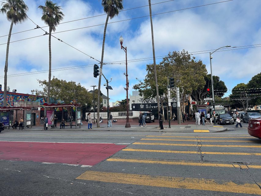 A city street intersection with pedestrians crossing, cars stopped at a red light, palm trees, and colorful murals on building walls.