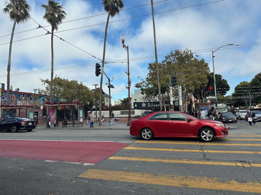 A red car waits at a crosswalk on an urban street corner with pedestrians, palm trees, traffic lights, and storefronts in the background.