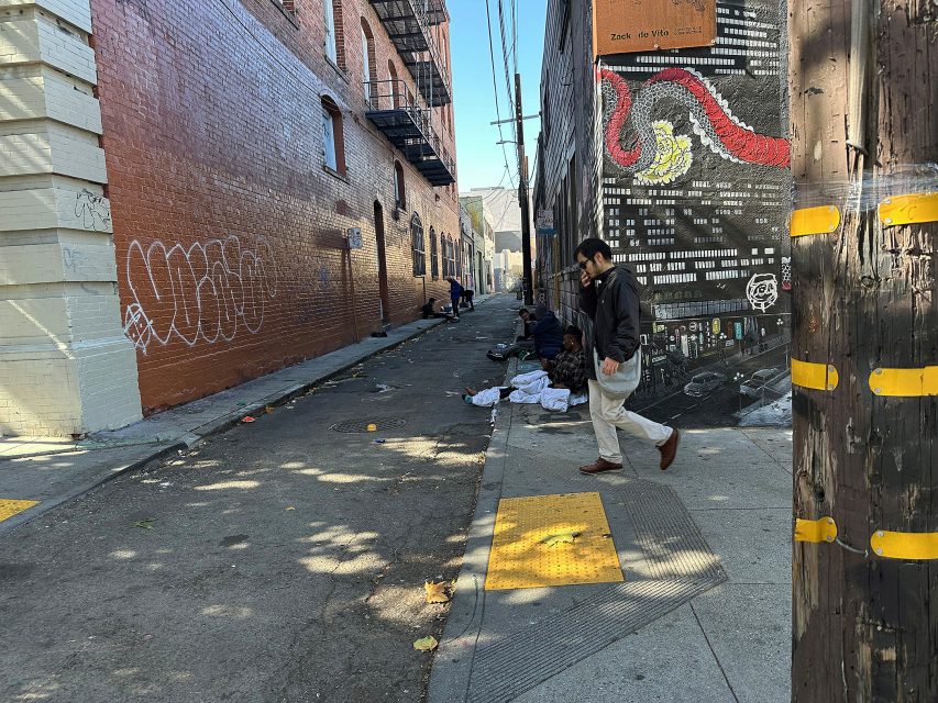 A man walks past an alley with scattered belongings and people sitting by a mural-covered wall in an urban setting.