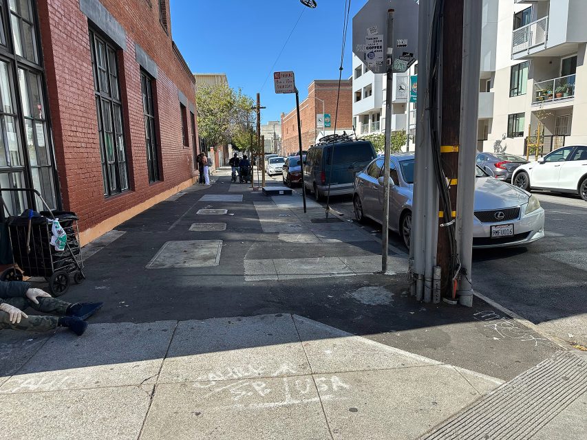 A city sidewalk with parked cars, a few people in the distance, a shopping cart, and some chalk writing on the pavement in an urban area.
