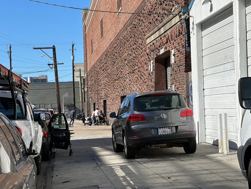 A gray Volkswagen SUV is parked near a brick building and white garage door; people are sitting on the sidewalk in the background on a sunny day.