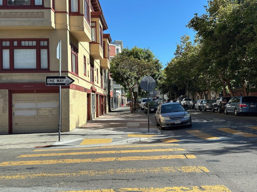 A city street with a one way sign, yellow crosswalk lines, parked cars, and apartment buildings under a clear blue sky.