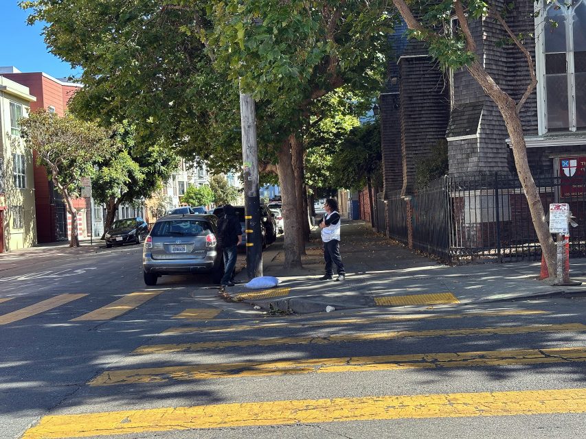 Two people stand on a sidewalk near a parked car and a tree on a sunny street. One person is taking a photo or video, and yellow crosswalk lines are visible in the foreground.