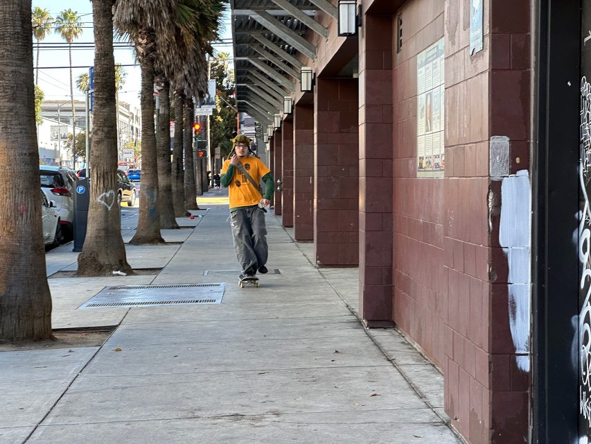 A person wearing a yellow shirt and green sweater skateboards down a city sidewalk lined with palm trees and brick buildings.