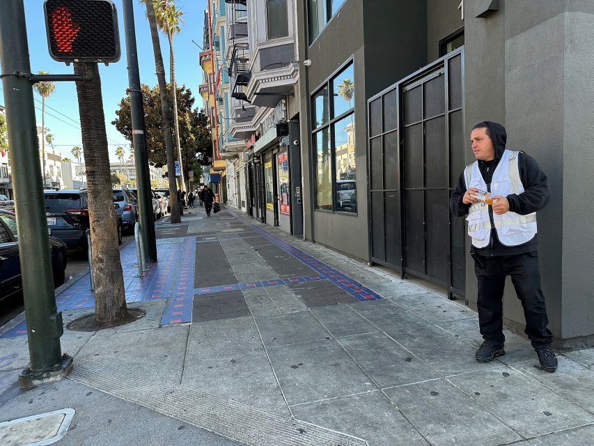 A man in a white vest stands on a city sidewalk holding food, near a building entrance and a traffic signal displaying a red hand.