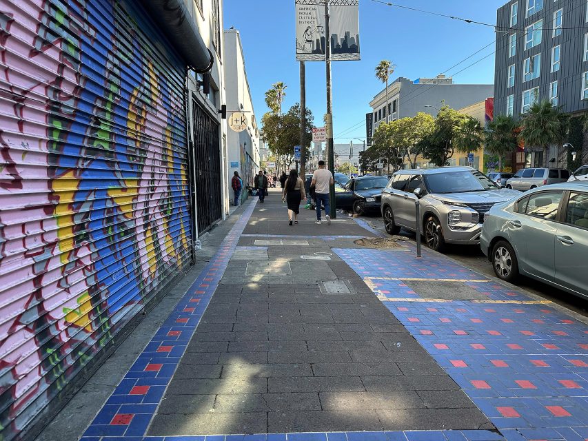 Pedestrians walk on a city sidewalk with colorful murals, blue and red tile sections, parked cars, and buildings lining the street on a sunny day.