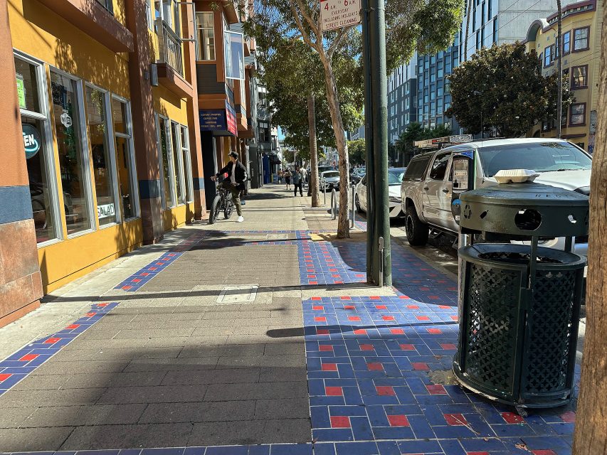 A city sidewalk with blue and red tile patterns, a trash can, parked cars, and people walking near buildings on a sunny day.
