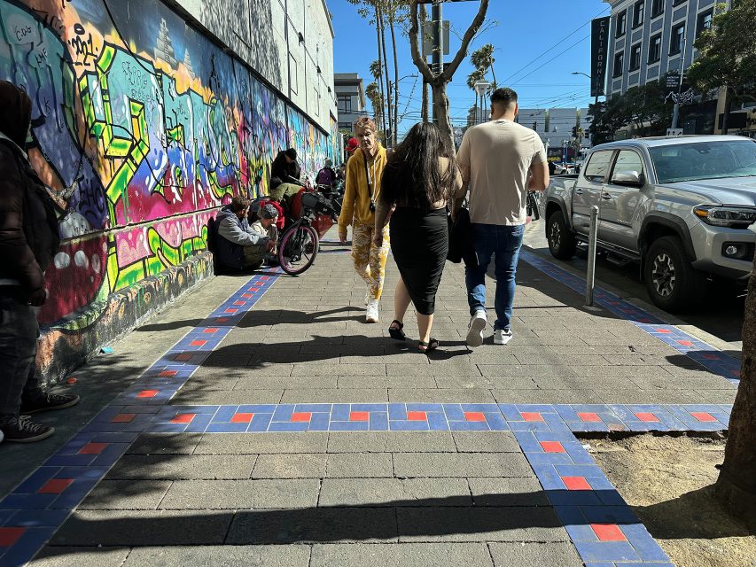 People walk on a sunlit city sidewalk with colorful graffiti on the wall, parked cars, and others sitting or standing nearby.