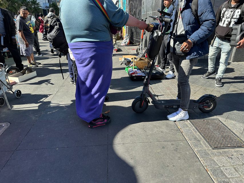 A group of people stand on a city sidewalk next to electric scooters, with street vendors and assorted items visible in the background.