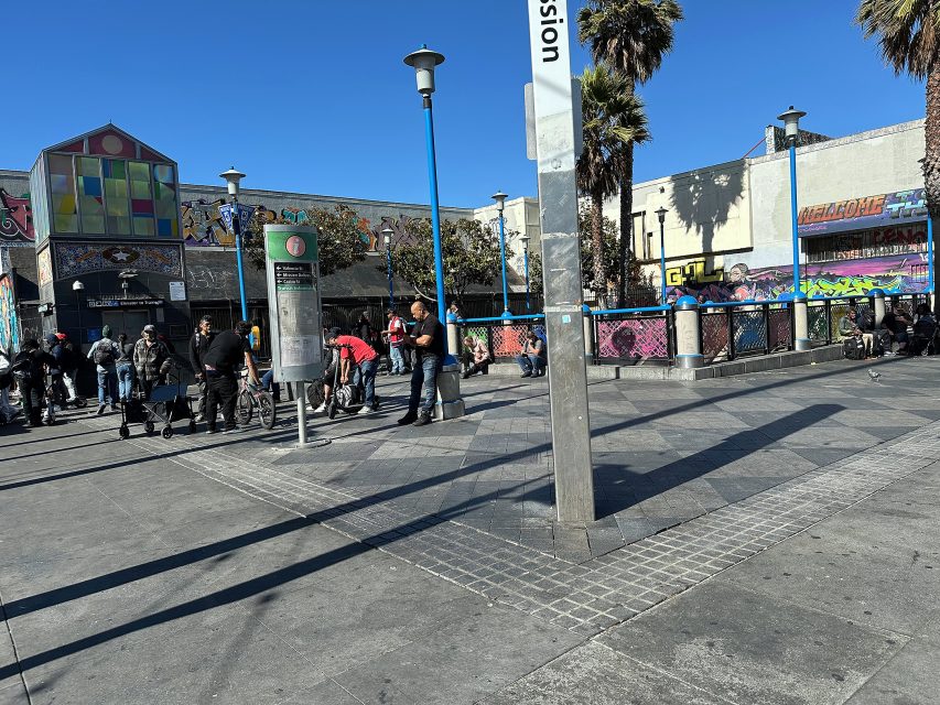 People stand and walk near a public payphone and mural-covered buildings on a sunny day in an urban outdoor plaza.