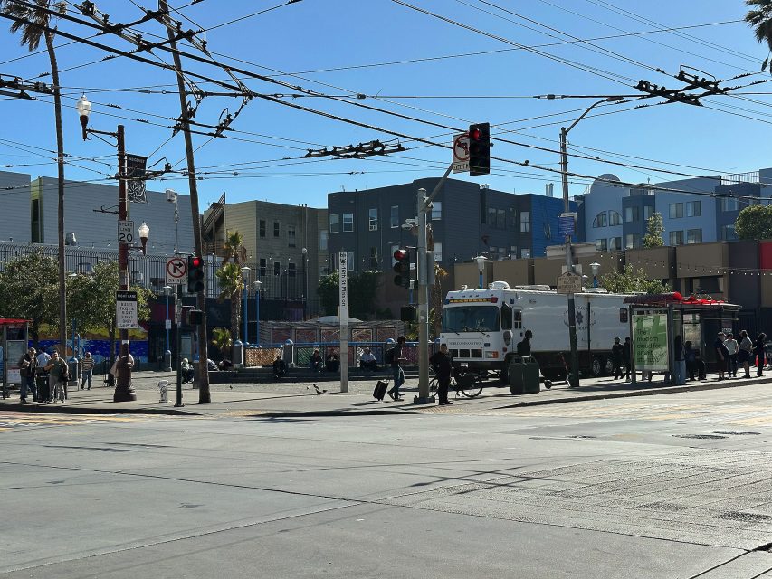 City street scene with people waiting at a bus stop, a large white utility vehicle, overhead power lines, and apartment buildings in the background under a clear sky.