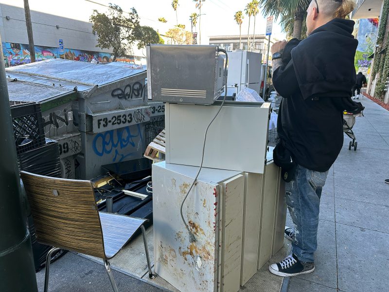 A person stands next to a stack of discarded appliances and furniture on a city sidewalk, with graffiti-covered dumpsters and a street in the background.