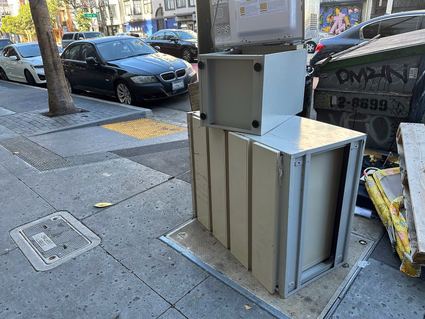 Several discarded metal cabinets and appliances are stacked on a city sidewalk next to a dumpster, with parked cars and buildings in the background.