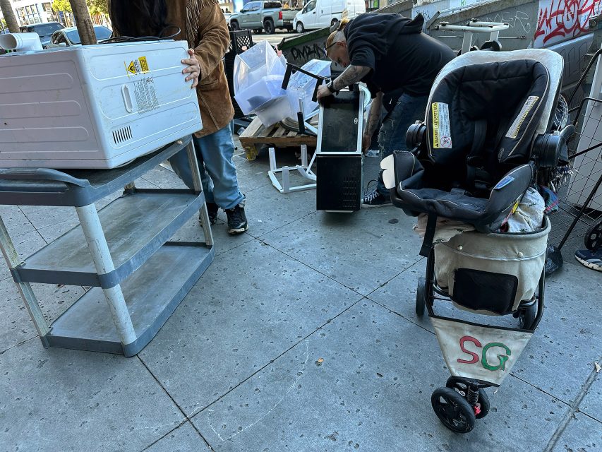 Two people sort through discarded household items on a city sidewalk; a stroller labeled "SG" is in the foreground.