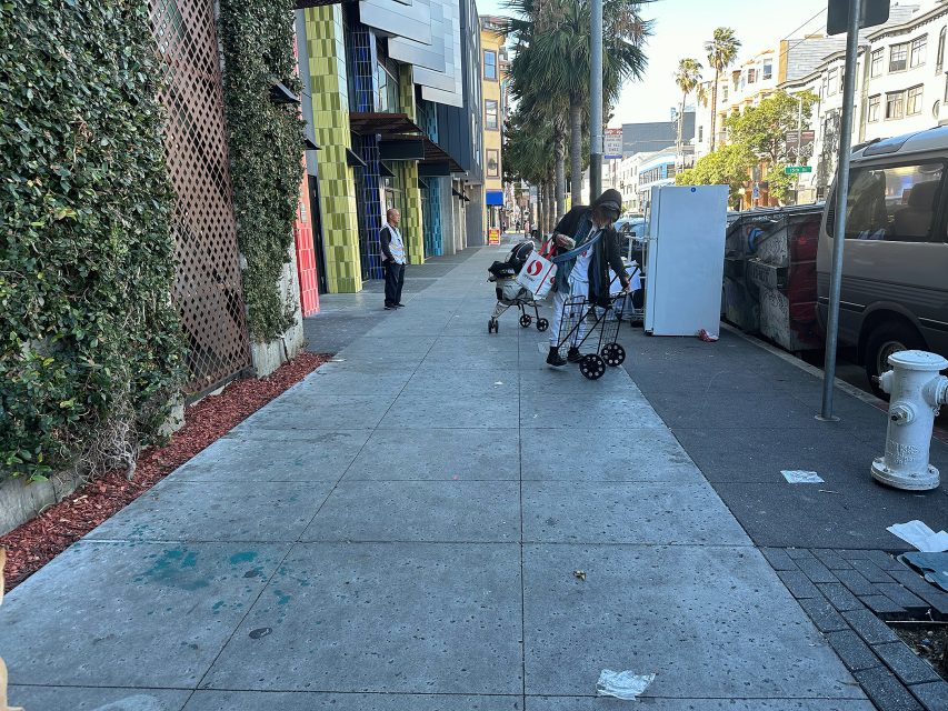 A person with carts sorts belongings on a city sidewalk; another person stands near a building, with cars parked along the street and a fire hydrant in view.