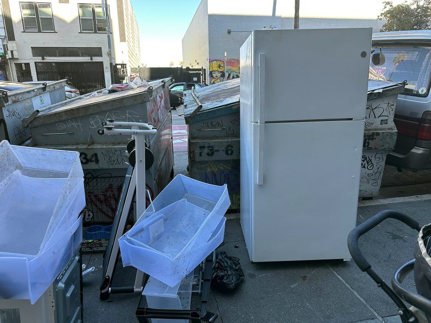 A white refrigerator and plastic storage bins are left on the sidewalk in front of graffiti-covered dumpsters on an urban street.