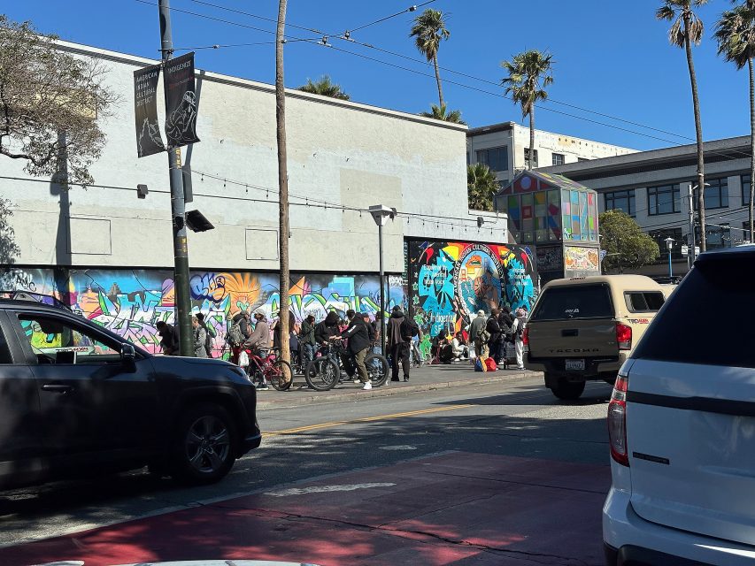 A group of people stands and sits near a colorful mural on the side of a building along a city street with cars parked and palm trees overhead.