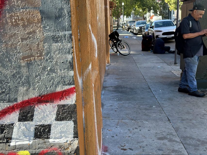 A man stands on a sidewalk near a textured wall with graffiti; in the background, two people sit on the ground and parked cars line the street.