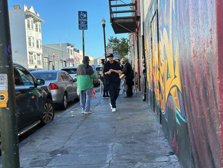 People walking and standing on a city sidewalk beside parked cars and a colorful graffiti mural during the daytime.