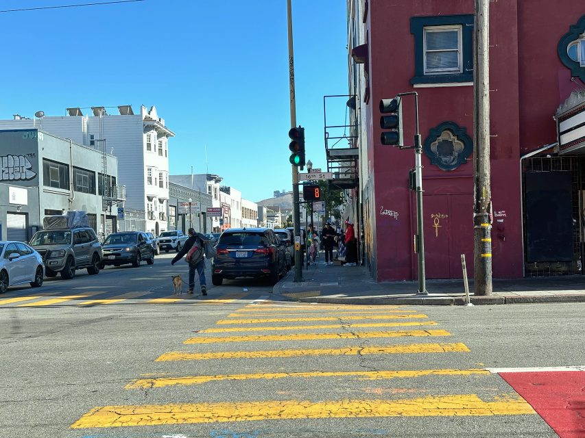 A city street intersection with yellow crosswalk lines, cars stopped at a red light, pedestrians waiting to cross, and buildings lining both sides of the road.