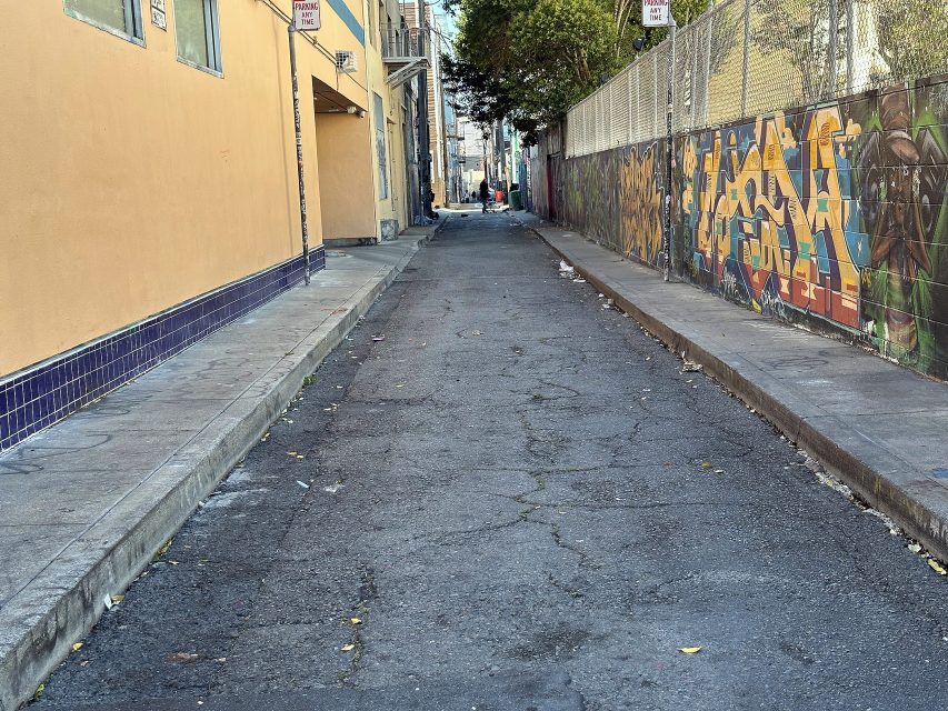 A narrow, cracked alleyway bordered by a yellow building on the left and a mural-covered fence on the right, with scattered litter on the ground.