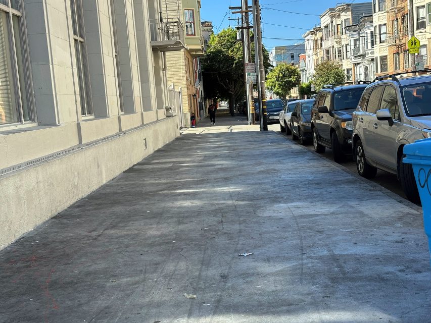 A sidewalk in a city lined with parked cars and multi-story buildings on a sunny day. A person is walking in the distance.