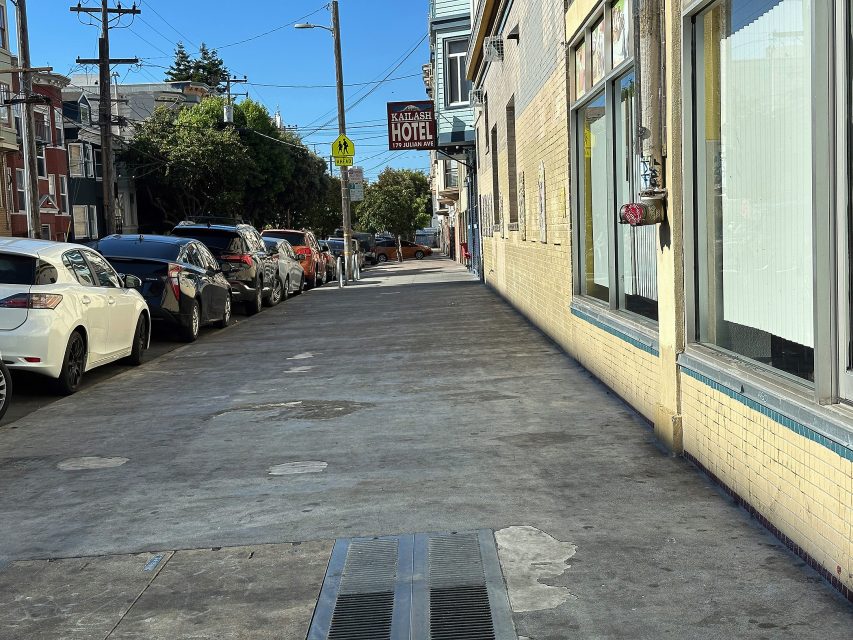 A city sidewalk with parked cars on the left and buildings on the right, including a hotel sign in the background under clear blue sky.