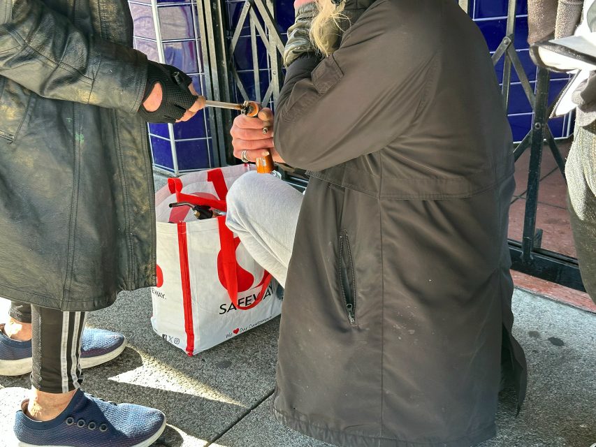 Two people interact on a sidewalk; one is kneeling with a beer bottle, and a white and red Safeway bag is on the ground between them.